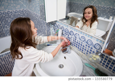 Young Girl Washing Hands at Bathroom Sink Reflected in Mirror, Cleanliness and Family Moment 133580779