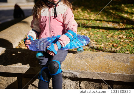 Kid With Blue Skateboard On Park Wall In Fall Light Wearing Pink Jacket And Knee Pads Kid With Blue Skateboard On Park Wall In Fall Light Wearing Pink Jacket And Knee Pads 133580823