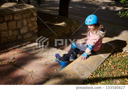 Young Girl With Blue Helmet Skates and Sits On Stone Wall In Park During Outdoor Play 133580827