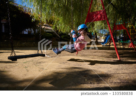 Girl in Pink Jacket Swinging at Playground With Helmet in Sunny Park 133580838