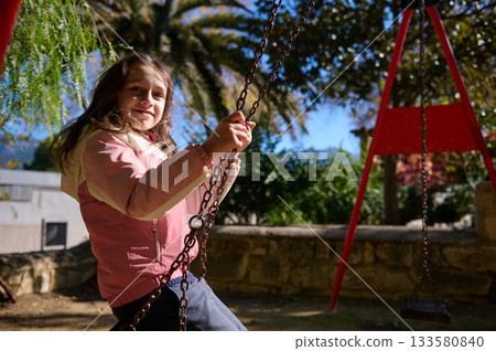 Joyful Girl On A Swing Outdoors, Sunlit Park Scene With Colorful Playground Equipment 133580840