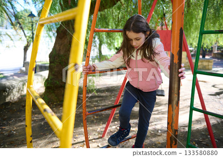 Young Girl Climbs Colorful Jungle Gym At Park, Exploring Outdoor Play Young Girl Climbs Colorful Jungle Gym At Park, Exploring Outdoor Play 133580880