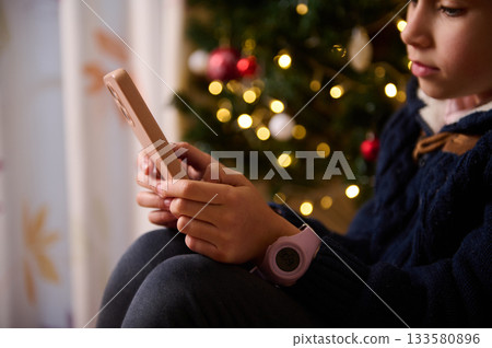 Child Using Smartphone Near Christmas Tree With Warm Lights And Cozy Holiday Mood 133580896