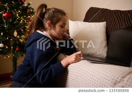 Young Girl Studying Next to Christmas Tree with Laptop at Home - Cozy Scene 133580898