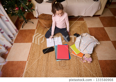 Little Girl Studying At Home On Rug With Notebooks, Christmas Tree And Teddy Bear Little Girl Studying At Home On Rug With Notebooks, Christmas Tree And Teddy Bear 133580908