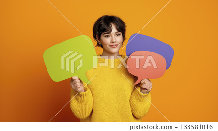 Young woman holding colorful speech bubble signs smiling with warm expression on orange background 133581016
