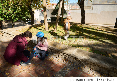 Family Helps Little Girl Tie Skates In Park While One Child Walks Nearby On Sunny Day Family Helps Little Girl Tie Skates In Park While One Child Walks Nearby On Sunny Day 133581028