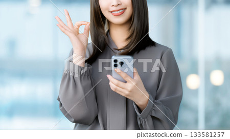 A smiling woman/businesswoman holding a smartphone and making an OK pose 133581257