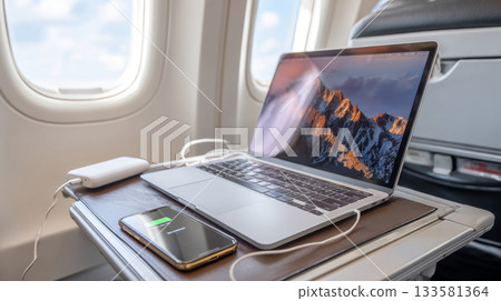 Laptop and smartphone charging on airplane tray table, showcasing remote work setup with power bank Laptop and smartphone charging on airplane tray table, showcasing remote work setup with power bank 133581364