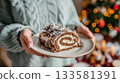 Woman in cozy sweater holds a slice of buche de noel on a plate, decorated Christmas tree softly blurred in the background, creating a festive atmosphere 133581391
