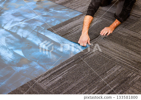 Carpenter is laying down carpet tiles in modern office setting, highlighting installation process. 133581800