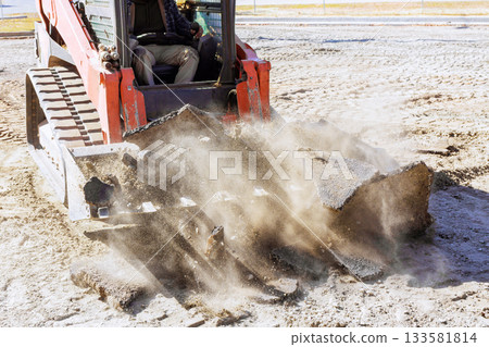 Construction worker uses skid steer loader to break up old asphalt in work area. 133581814