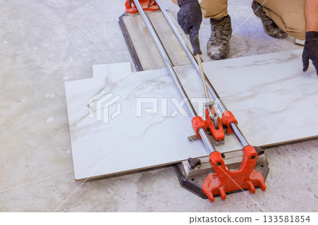 Worker uses tile cutter to slice large marble tiles for flooring renovation in contemporary room. Worker uses tile cutter to slice large marble tiles for flooring renovation in contemporary room. 133581854