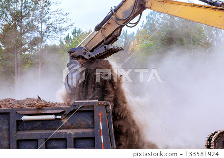 Construction equipment transfers dirt debris into large container while dust clouds rise in air. 133581924
