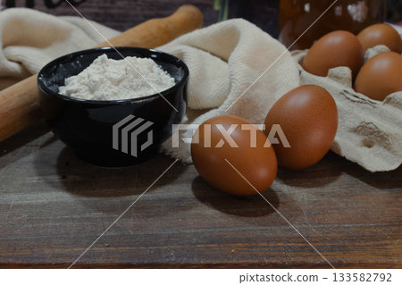 Baking ingredients on a rustic wooden board, showing eggs in a carton, flour in a black bowl, and a rolling pin for preparing homemade pastry recipes Baking ingredients on a rustic wooden board, showing eggs in a carton, flour in a black bowl, and a rolling pin for preparing homemade pastry recipes 133582792