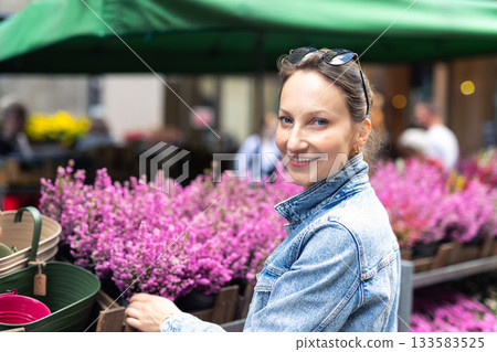 Young adult joyful woman choose to buy colorful potted flowers at outdoor market stall. Female person at sale of blooming plants in pots at botanical street German store on sunny spring autumn day 133583525