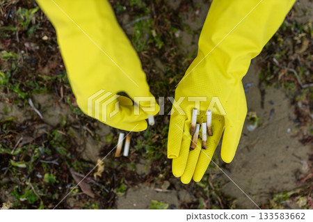 Close-up of a volunteer wearing yellow gloves collecting cigarette butts on a polluted beach. Environmental cleanup, ecology protection, shoreline waste and anti-litter awareness. Close-up of a volunteer wearing yellow gloves collecting cigarette butts on a polluted beach. Environmental cleanup, ecology protection, shoreline waste and anti-litter awareness. 133583662