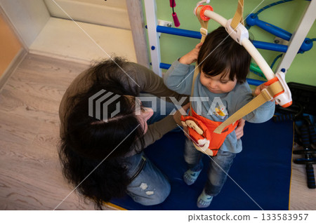 Child engaging in playful movement with support harness, while caregiver encourages exploration during activity session in a cheerful indoor play area 133583957