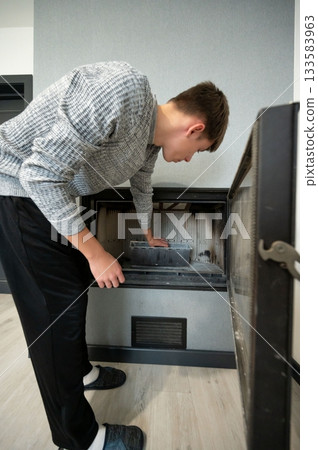 Young man carefully cleaning the inside of a sleek fireplace in a modern living room, focusing on removing ash and debris for a tidy appearance 133583963