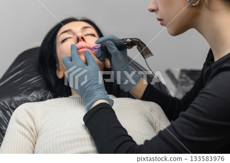 Close up of a cosmetologist performing permanent lip makeup, tattooing natural lip color enhancement with sterile tools in a professional beauty studio. Close up of a cosmetologist performing permanent lip makeup, tattooing natural lip color enhancement with sterile tools in a professional beauty studio. 133583976