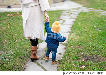 First steps into fall a tender moment on a stone path shared between a mother in boots and her toddler reaching up with curiosity and joy, autumn time exploration 133584094