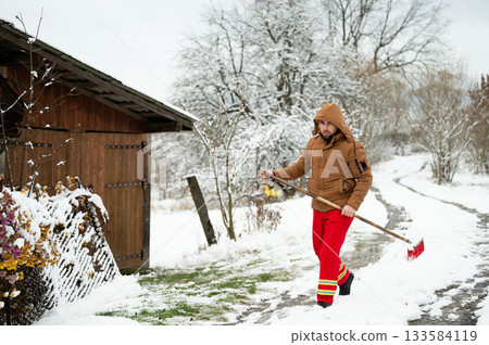 Snowy winter day brings a worker outside to clear the path near a rustic cabin surrounded by trees blanketed in white 133584119