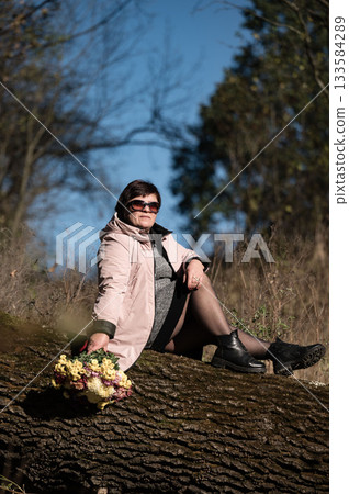 Woman relaxing on a log in a serene natural setting on a sunny day, enjoying the beauty of nature with a bouquet of flowers in hand 133584289