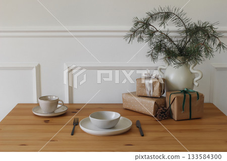 Minimalist holiday table setting with ceramic plate, fork cutlery. Wrapped Gifts, pine tree branch decor on wooden table. Cup of tea, coffee. Christmas interior styling. Seasonal winter home design 133584300