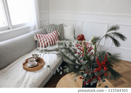 Cozy Christmas living room interior. Pine, red holly berry floral arrangement, Festive holiday home decor. Blurred background. Scandinavian style sofa with pillows, window. Winter morning coffee scene 133584305