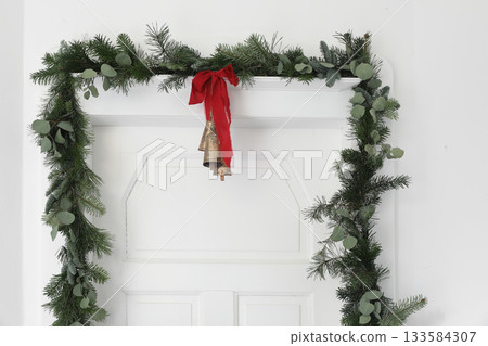 Christmas door garland with fir, pine tree branches, eucalyptus, red ribbon. Festive winter doorway decoration with vintage bells, natural greenery, Holiday home styling. Empty white wall copy space. 133584307