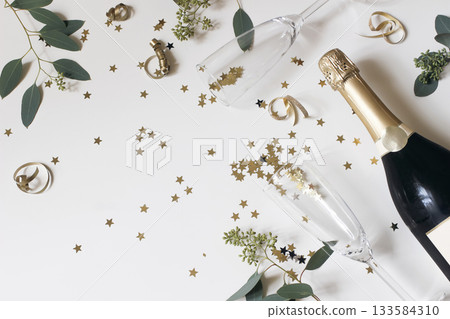 Happy New Year still life. Empty champagne glasses, wine bottle. Golden star confetti, eucalyptus tree branches isolated on white table background. Christmas celebration, party. Flat lay, top view. 133584310