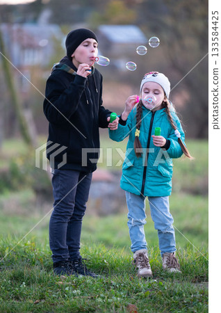 Two children play joyfully in a lush green field on a crisp autumn afternoon, blowing bubbles and enjoying each other's company as laughter fills the air 133584425