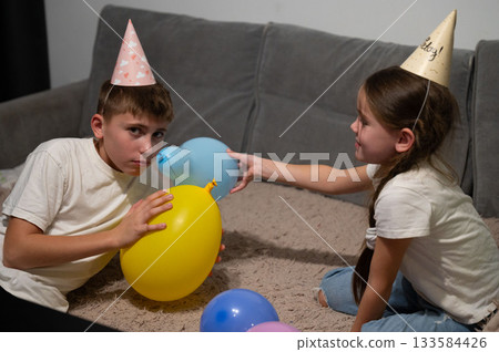 Celebrating joy and laughter with balloons and party hats, two children engage in playful fun at a cozy indoor setting during an exciting birthday gathering Celebrating joy and laughter with balloons and party hats, two children engage in playful fun at a cozy indoor setting during an exciting birthday gathering 133584426
