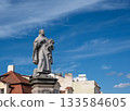 Prague, czech republic, august 9, 2023. Stone statue of a saint wearing robes and holding flowers, standing on a pedestal with an ornate base against a bright blue sky 133584605