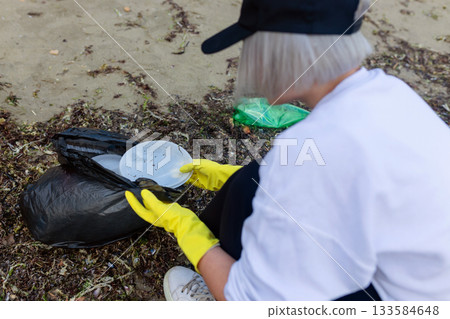 Volunteer collecting plastic waste and litter into a black trash bag during beach cleanup, highlighting pollution, environmental protection, sustainability, and eco awareness. Volunteer collecting plastic waste and litter into a black trash bag during beach cleanup, highlighting pollution, environmental protection, sustainability, and eco awareness. 133584648