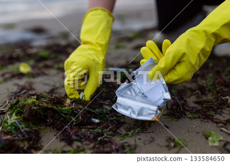 Hands in yellow gloves picking up crushed packaging and litter during beach cleanup, showing pollution, environmental damage and the importance of eco awareness and coastal protection. 133584650
