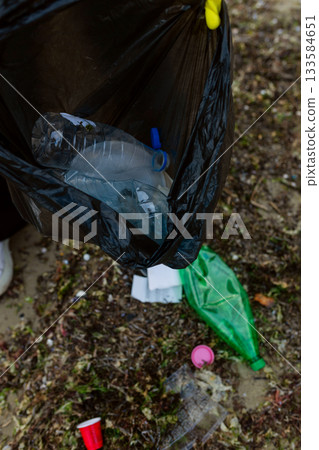 Volunteer collecting plastic waste and litter into a black trash bag during beach cleanup, highlighting pollution, environmental protection, sustainability, and eco awareness. 133584651