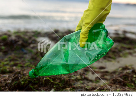 Volunteer picking up a green plastic bottle during beach cleanup, highlighting pollution, waste removal, and environmental protection. 133584652