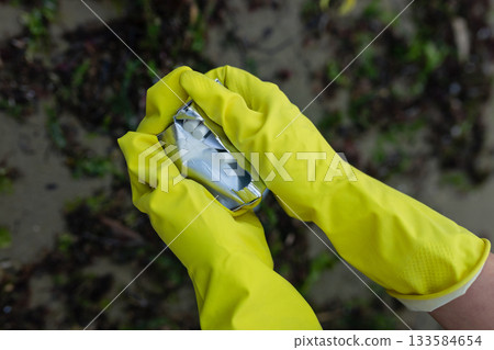 Hands in yellow gloves picking up crushed packaging and litter during beach cleanup, showing pollution, environmental damage and the importance of eco awareness and coastal protection. Hands in yellow gloves picking up crushed packaging and litter during beach cleanup, showing pollution, environmental damage and the importance of eco awareness and coastal protection. 133584654