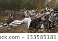 pile of corrugated sheets and debris scattered near forest edge; rusted metal, broken windows, wooden pallets and plastic bags create neglected backyard eyesore with overgrown grass 133585181