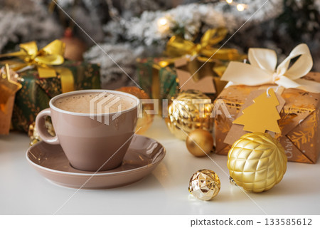 Cup of Hot Drink with Christmas Gifts and Ornaments in Cozy Holiday Setting against Christmas Tree and Lights 133585612