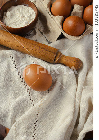 A top-down view of freshly baked ingredients laid out on a linen cloth, a whole brown egg, bowls of flour, and a rolling pin, ready for making homemade pastries and comforting recipes. A top-down view of freshly baked ingredients laid out on a linen cloth, a whole brown egg, bowls of flour, and a rolling pin, ready for making homemade pastries and comforting recipes. 133585745