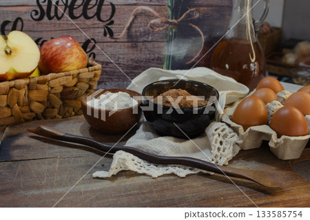 High-quality baking ingredients like fresh apples, cocoa powder, white flour, and brown eggs arranged with a dark wooden spoon highlighting home cooking and sweet food preparation 133585754