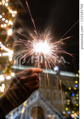 hand holding sparkler in front of Christmas tree. picture shows festive and joyful mood. 133585789