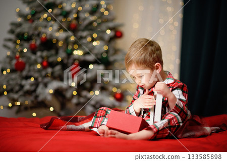child in festive plaid pajamas opening a present while sitting near a beautifully lit Christmas tree. Warm holiday atmosphere with soft bokeh lights and cozy seasonal colors. child in festive plaid pajamas opening a present while sitting near a beautifully lit Christmas tree. Warm holiday atmosphere with soft bokeh lights and cozy seasonal colors. 133585898