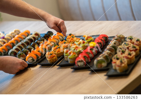 A person hands arrange multiple large, gourmet platters of diverse sushi rolls 133586263