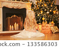 Happy little girl child with festive gift box sitting near Christmas tree and fireplace at home 133586303
