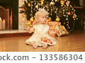Happy little girl child opens festive gift box sitting near Christmas tree and fireplace at home 133586304