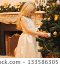 Happy little girl child with festive gift box sitting near Christmas tree and fireplace at home 133586305