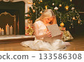 Happy little girl child with festive gift box sitting near Christmas tree and fireplace at home 133586308